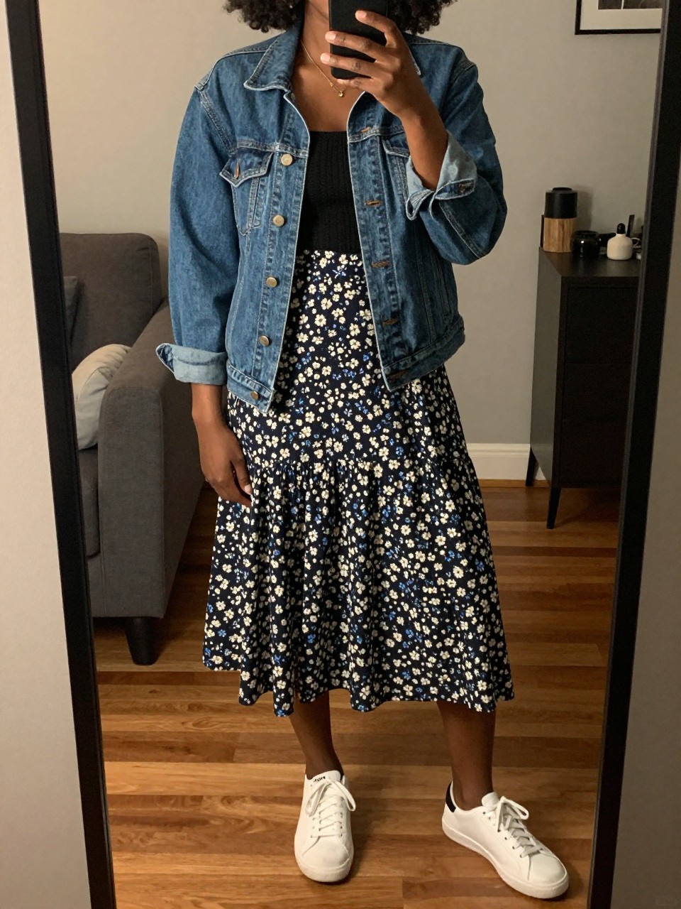 iPhone mirror selfie of a black woman in a denim jacket and a floral cotton skirt with white sneakers, one hand resting on her hip, cropped at NECK so face doesn't show, in a city apartment, warm moody lighting, iPhone photo quality.