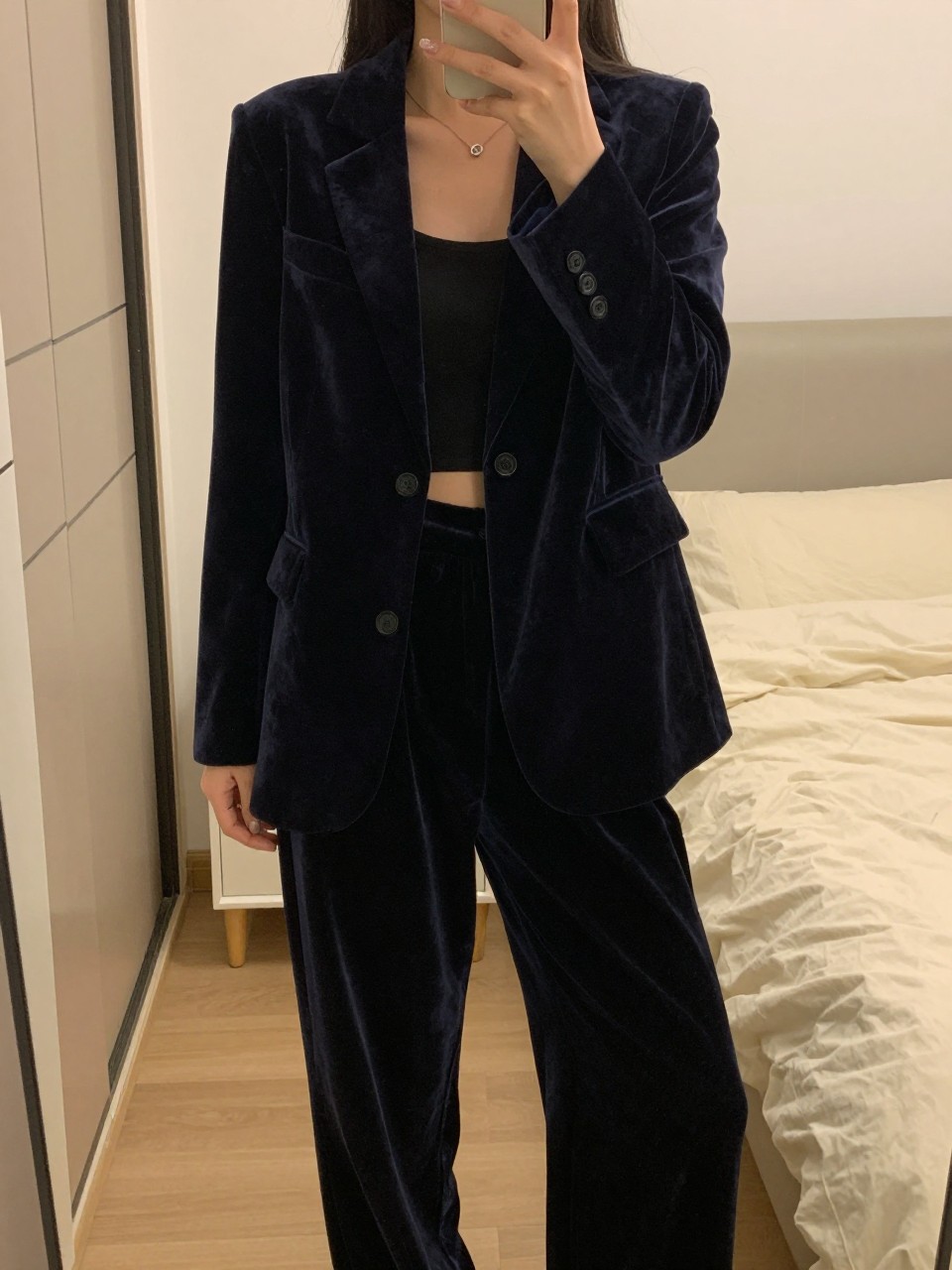 mirror selfie of a woman in a velvet blazer and wide-leg pants, blazer worn open, cropped at NECK so face doesn't show, in a feminine-themed bedroom, warm moody lighting, iPhone photo quality.