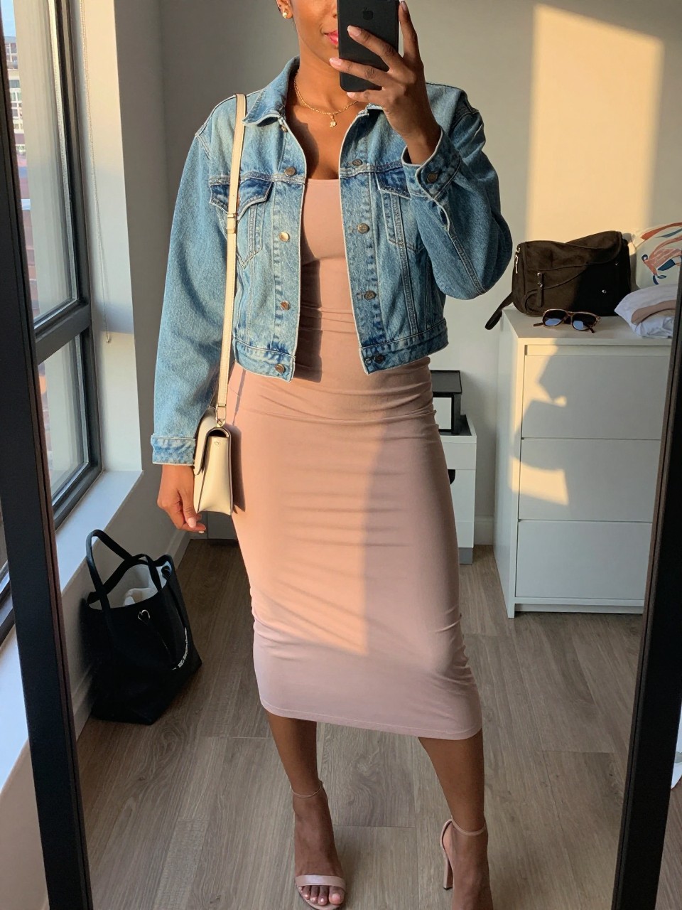 mirror selfie of a black woman wearing a blush column midi dress with a light wash cropped denim jacket, nude heeled sandals, a cream mini crossbody bag, and soft pink lipstick, holding a phone in front of a mirror, cropped at NECK so face doesn't show, with a natural casual selfie pose, in a city apartment, evening golden hour, including small lived-in details like a tote bag on the floor, sunglasses on a dresser, or soft bedding in the background, latest iPhone photo quality.
