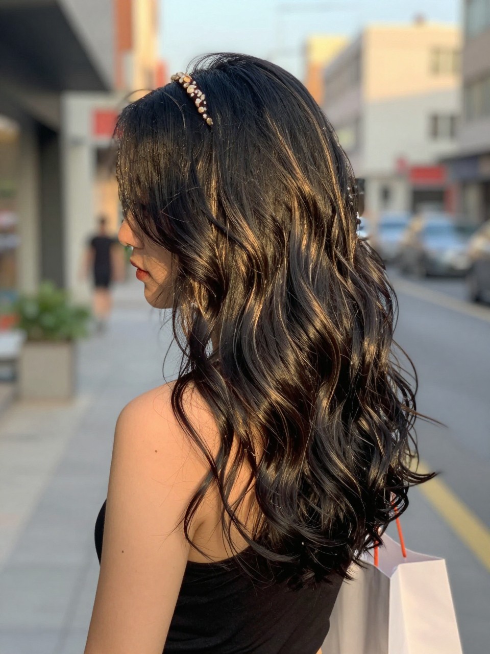 Photo of a woman wearing beachy black waves with sunlit highlight streaks and soft texture, side profile view, in a softly blurred city street background, golden hour glow, containing a small lived-in detail such as a headband pushed back and a few shopping bags nearby, latest iPhone photo quality.