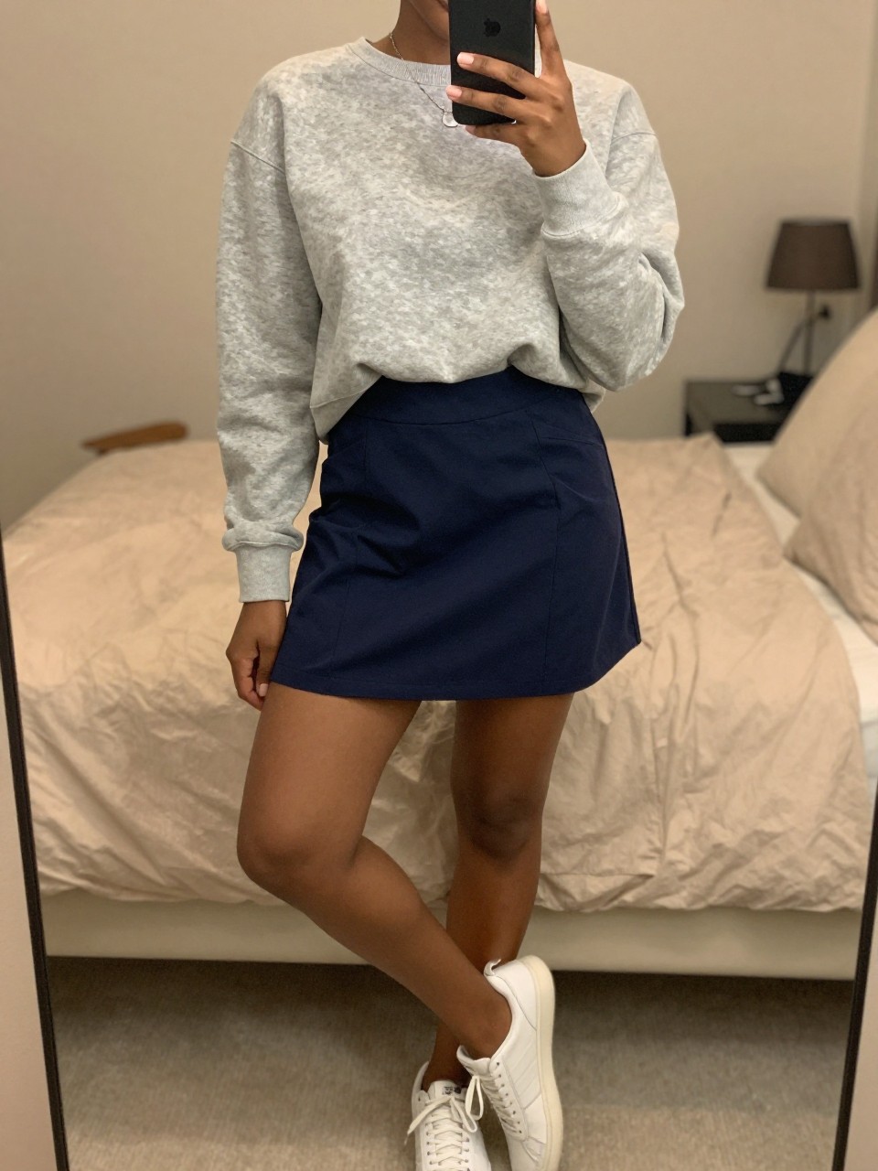 iPhone mirror selfie of a black woman wearing a heathered gray sweatshirt half-tucked into a navy structured cotton skirt with white sneakers, holding her phone low by her thigh, cropped at NECK so face doesn't show, in a feminine-themed bedroom, warm moody lighting, iPhone photo quality.