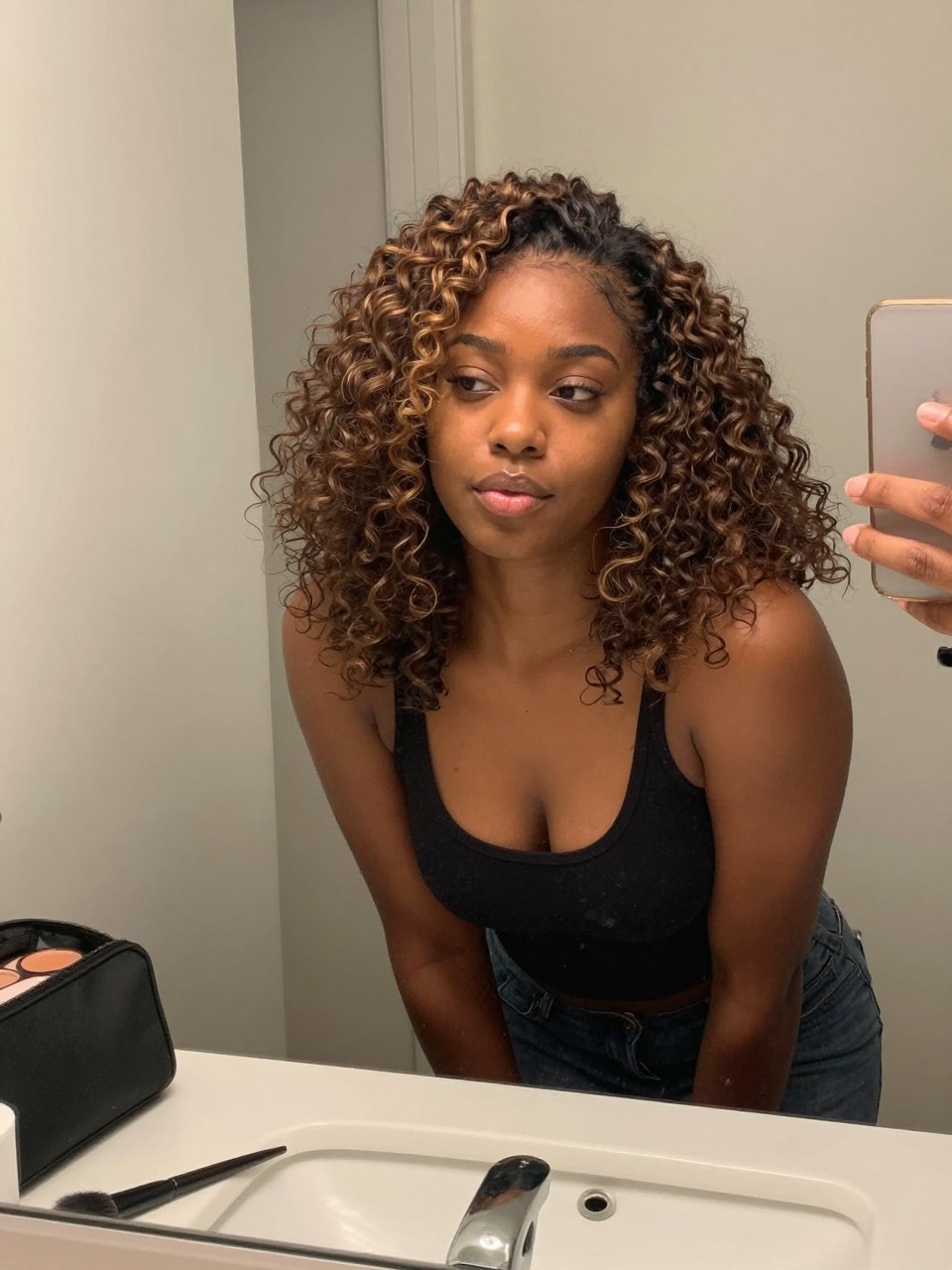 Photo of a Black woman wearing a natural wash and go with caramel highlights glowing through defined curls, candid mirror selfie, at a bathroom vanity mirror, soft indoor lighting, containing a small lived-in detail such as a defining brush on the counter and a makeup bag nearby, latest iPhone photo quality.