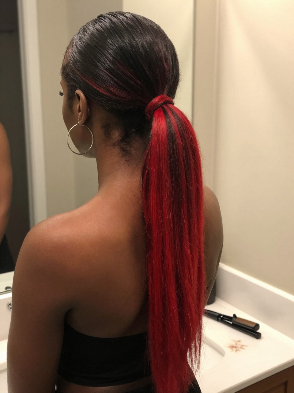 Photo of a black woman wearing a sleek mid ponytail with black hair and bright red highlight panels through the tail, back-of-head view to highlight details, at a bathroom vanity mirror, soft indoor lighting, containing a small lived-in detail such as a flat iron on the counter and hoop earrings peeking through, latest iPhone photo quality.