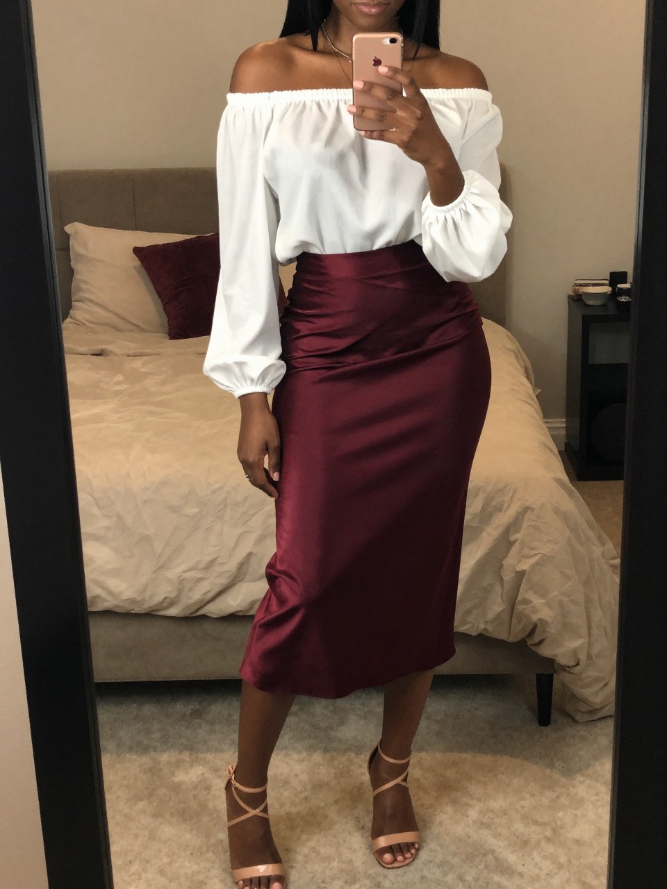 mirror selfie of a black woman wearing a white off-the-shoulder blouse tucked into a deep burgundy satin midi skirt with nude strappy block-heel sandals, one hand holding her phone at chest level, cropped at NECK so face doesn't show, in a feminine-themed bedroom, warm moody lighting, iPhone photo quality.