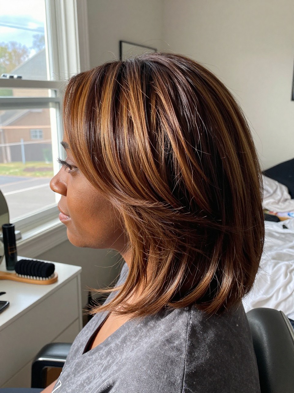 Photo of a black woman wearing a side part brown blowout with cinnamon highlights and deeper lowlights, side profile view, in a sun-drenched bedroom, natural window light, containing a small lived-in detail such as velcro rollers on the vanity and a round brush nearby, latest iPhone photo quality.