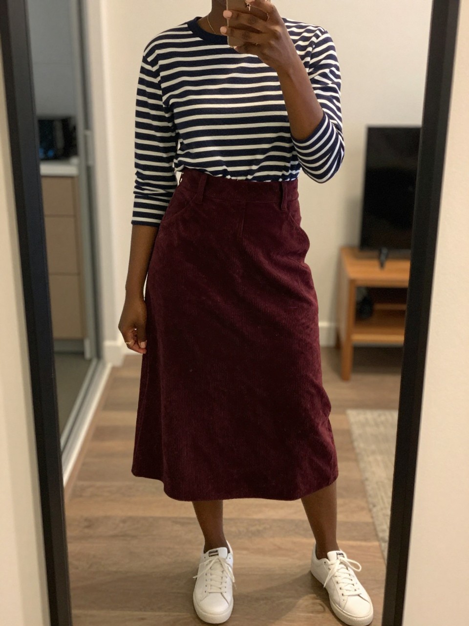 mirror selfie of a black woman wearing a navy striped Breton top tucked into a deep burgundy A-line corduroy skirt with white sneakers, one hand holding her phone at her side, cropped at NECK so face doesn't show, in a city apartment, warm moody lighting, iPhone photo quality.