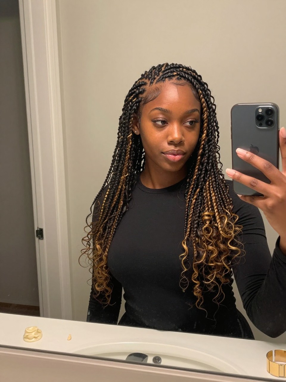 Photo of a Black woman wearing neat mini twists with golden brown highlights scattered throughout, candid mirror selfie, at a bathroom vanity mirror, soft indoor lighting, containing a small lived-in detail such as twisting cream on the counter and gold hair cuffs nearby, latest iPhone photo quality.