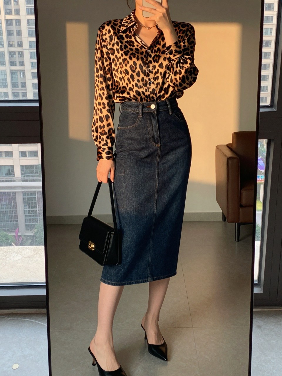 iPhone mirror selfie of a woman wearing a leopard print silk blouse tucked into a dark-wash denim pencil skirt with black heeled mules, holding a small black shoulder bag, standing in a confident pose, cropped at NECK so face doesn't show, in a city apartment, evening golden hour, iPhone photo quality.