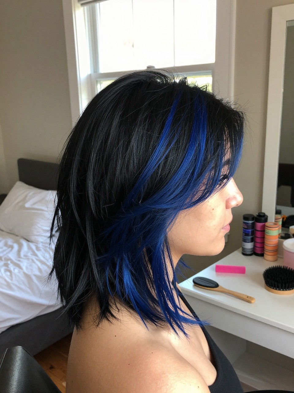 Photo of a woman wearing a side part black blowout with deep ocean blue highlights, side profile view, in a sun-drenched bedroom, natural window light, containing a small lived-in detail such as velcro rollers on the vanity and a round brush nearby, latest iPhone photo quality.