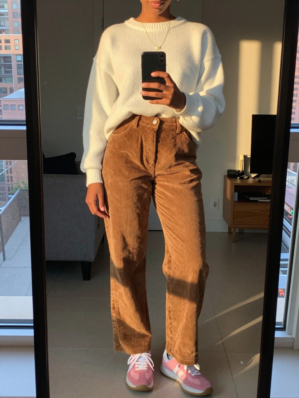 mirror selfie of a black woman in corduroy pants and a white sweater with pink sneakers, standing with phone at waist, cropped at NECK so face doesn't show, in a city apartment, evening golden hour, iPhone photo quality.