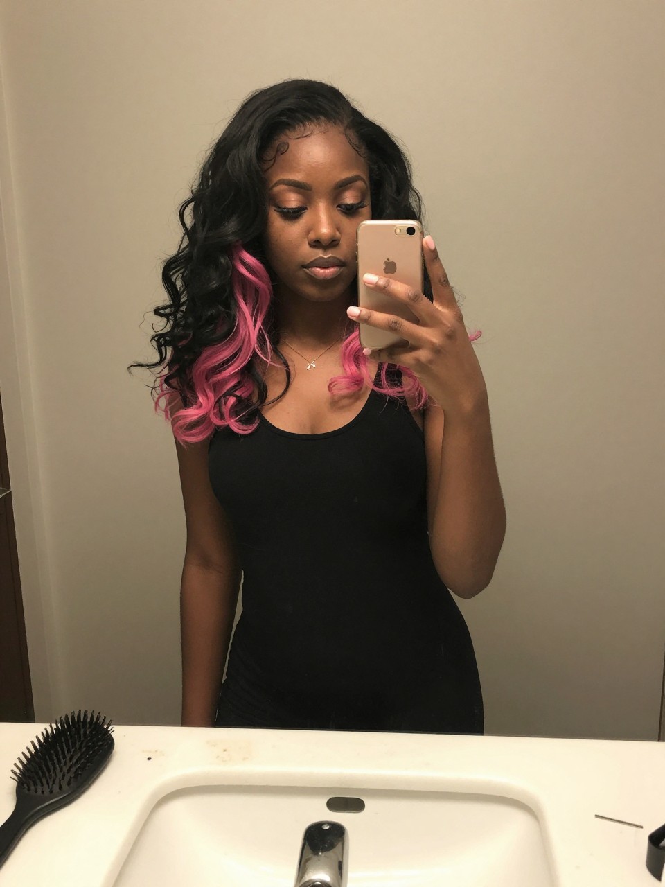 Photo of a black woman wearing a wavy black lob with bubblegum pink highlights, candid mirror selfie, at a bathroom vanity mirror, soft indoor lighting, containing a small lived-in detail such as a hairbrush on the sink and a silver clip nearby, latest iPhone photo quality.