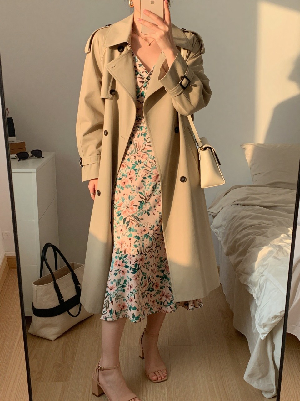 mirror selfie of a woman wearing a blush and green floral wrap midi dress with a warm beige light trench coat, nude block-heel sandals, medium gold hoop earrings, and a cream mini shoulder bag, holding a phone in front of a mirror, cropped at NECK so face doesn't show, with a natural casual selfie pose, in a feminine-themed bedroom, evening golden hour, including small lived-in details like a tote bag on the floor, sunglasses on a dresser, or soft bedding in the background, latest iPhone photo quality.