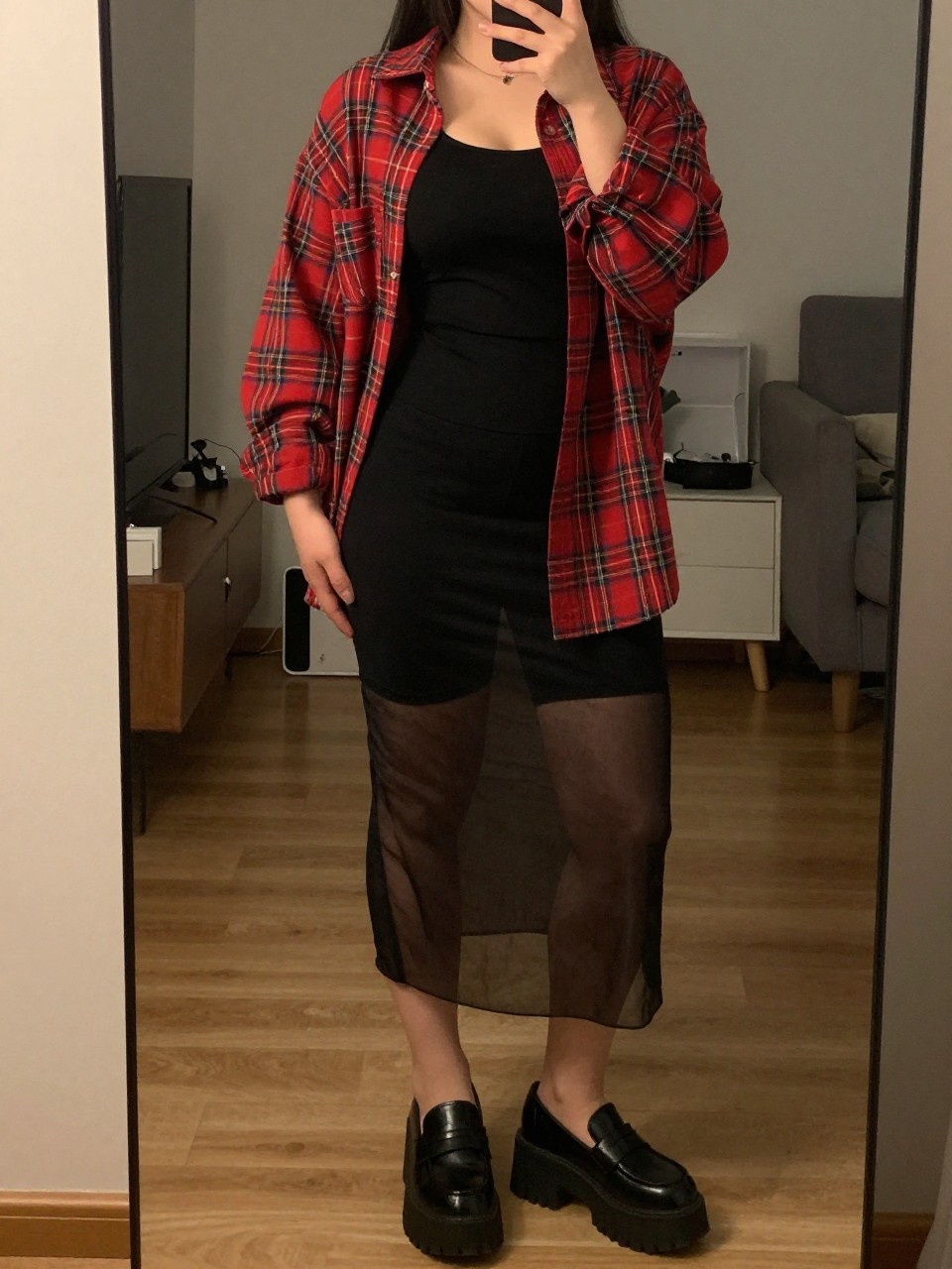 mirror selfie of curvy woman wearing an oversized red plaid shirt open over a black tank top with black bike shorts underneath a black sheer mesh skirt and chunky platform loafers, one hand in her pocket, cropped at NECK so face doesn't show, in a city apartment, warm moody lighting, iPhone photo quality.