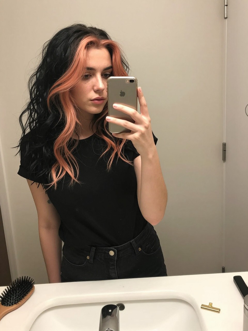 Photo of a woman wearing a wavy black lob with peach orange highlights, candid mirror selfie, at a bathroom vanity mirror, soft indoor lighting, containing a small lived-in detail such as a hairbrush on the sink and a gold clip nearby, latest iPhone photo quality.