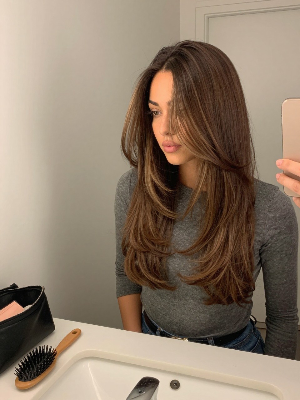 Photo of a woman wearing a sleek walnut brown lob with deep mocha highlights, candid mirror selfie, at a bathroom vanity mirror, soft indoor lighting, containing a small lived-in detail such as a hairbrush on the vanity and a makeup bag nearby, latest iPhone photo quality.