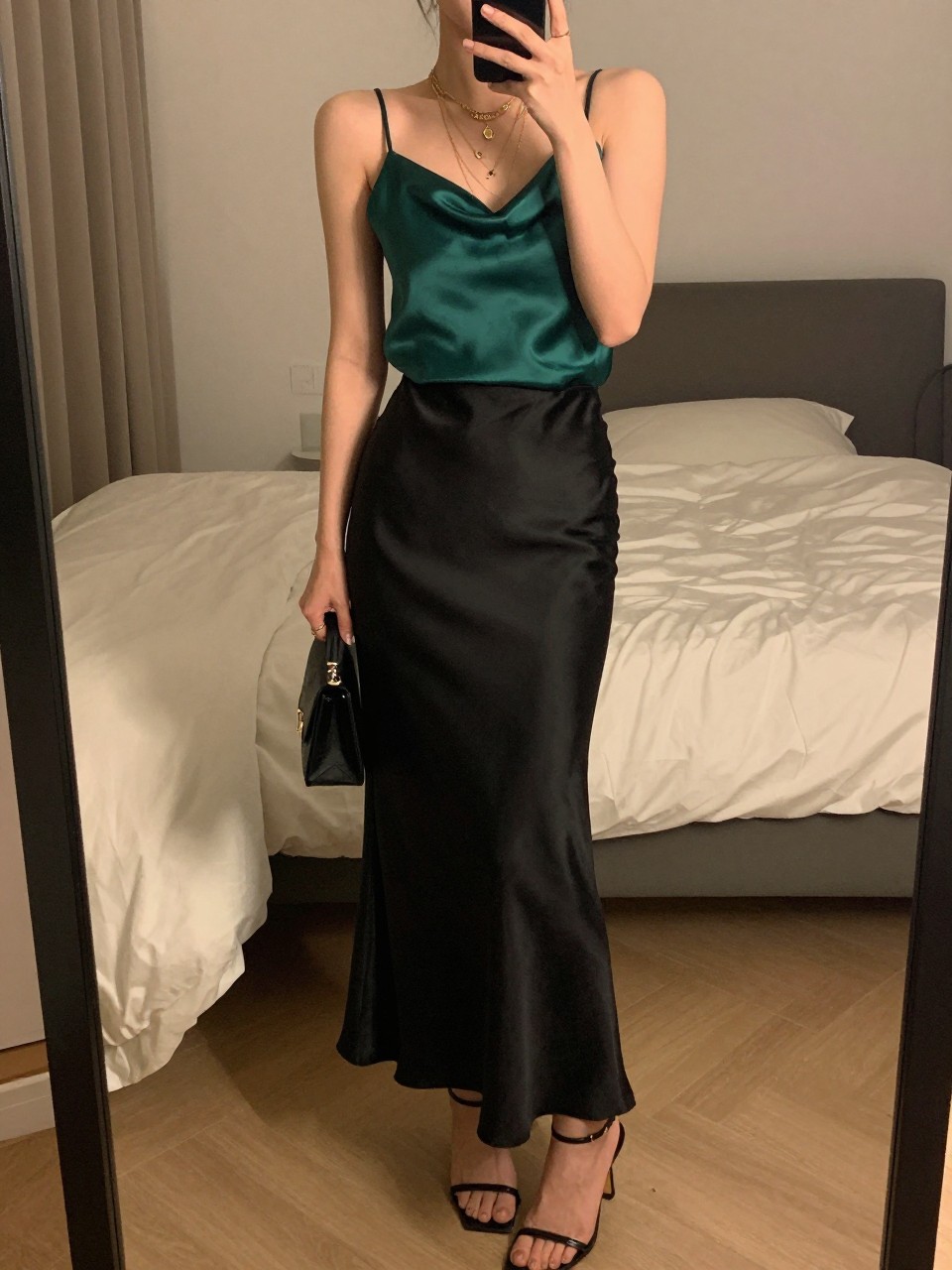 iPhone mirror selfie of a woman wearing an emerald silk camisole tucked into a black satin maxi skirt with black strappy heeled sandals and layered gold necklaces, holding a small black clutch, standing in a romantic evening pose, cropped at NECK so face doesn't show, in a feminine-themed bedroom, warm moody lighting, iPhone photo quality.