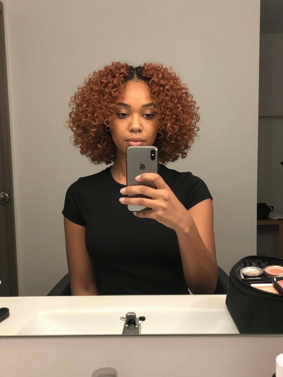 Photo of a black woman wearing an auburn brown curly bob with soft defined curls, candid mirror selfie, bathroom vanity mirror setting, soft indoor lighting, with small claw clips on the counter and a makeup bag nearby, latest iPhone photo quality.