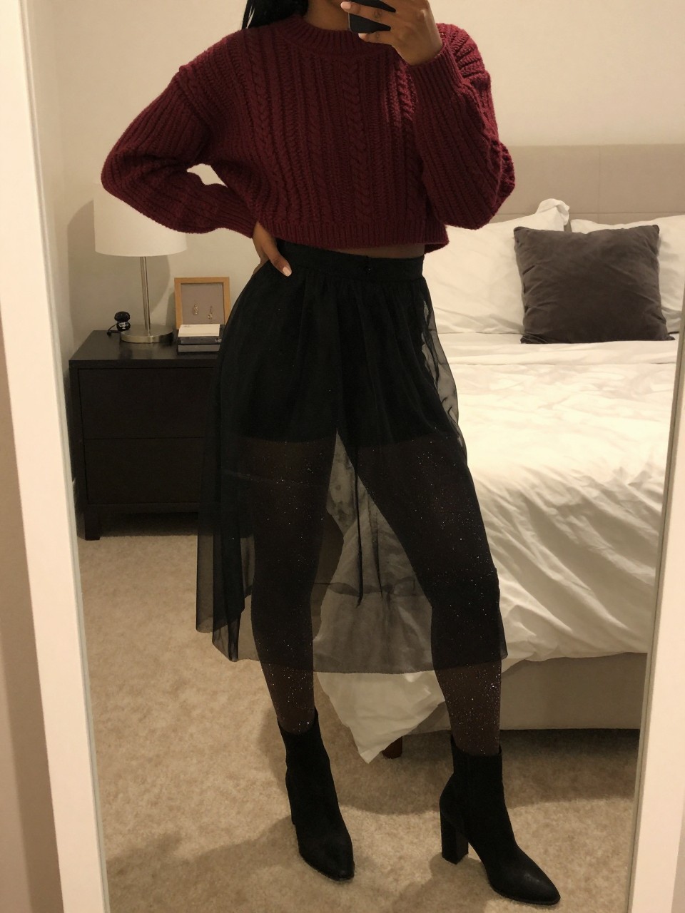mirror selfie of a black woman wearing a burgundy cropped cable-knit sweater with sparkly tights underneath a black sheer mesh skirt and black block-heel boots, one hand on her hip, cropped at NECK so face doesn't show, in a feminine-themed bedroom, warm moody lighting, iPhone photo quality.