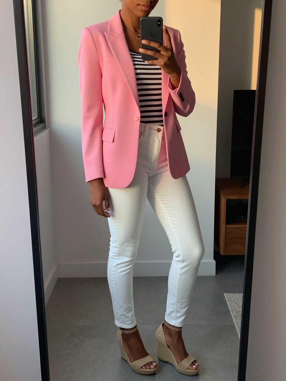 mirror selfie of a black woman in a pink blazer and striped top with white jeans and wedges, standing sideways looking at phone screen, cropped at NECK so face doesn't show, in a city apartment, evening golden hour, iPhone photo quality.