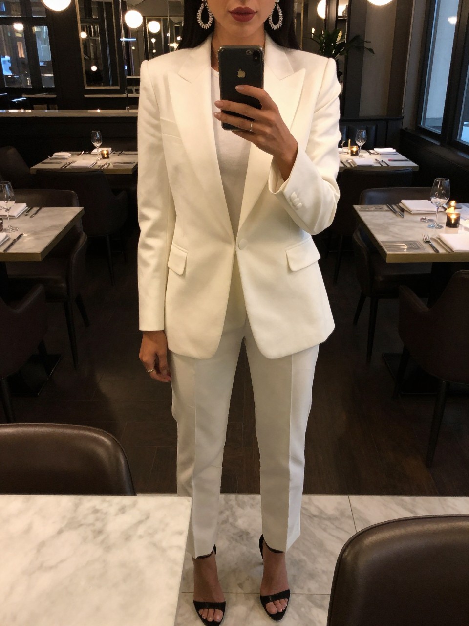iPhone mirror selfie of a woman in a white tuxedo set and statement earrings with heels, standing in a restaurant or bar, cropped at NECK so face doesn't show, in a city apartment, warm moody lighting, iPhone photo quality.
