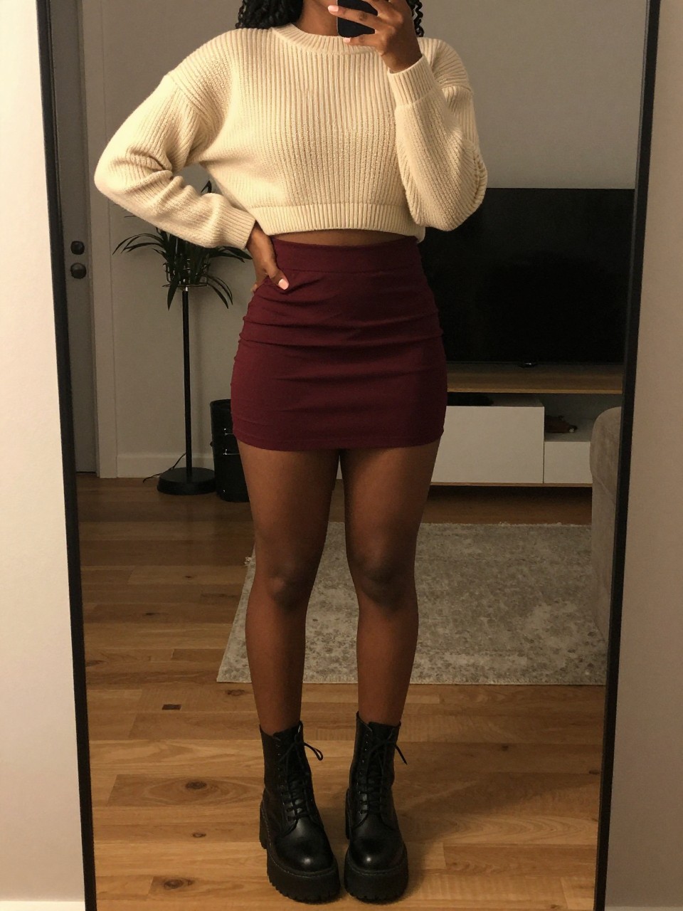 mirror selfie of a black woman wearing a cream cropped ribbed sweater with a high-waisted deep burgundy mini skirt and black platform combat boots, one hand on her hip, cropped at NECK so face doesn't show, in a city apartment, warm moody lighting, iPhone photo quality.