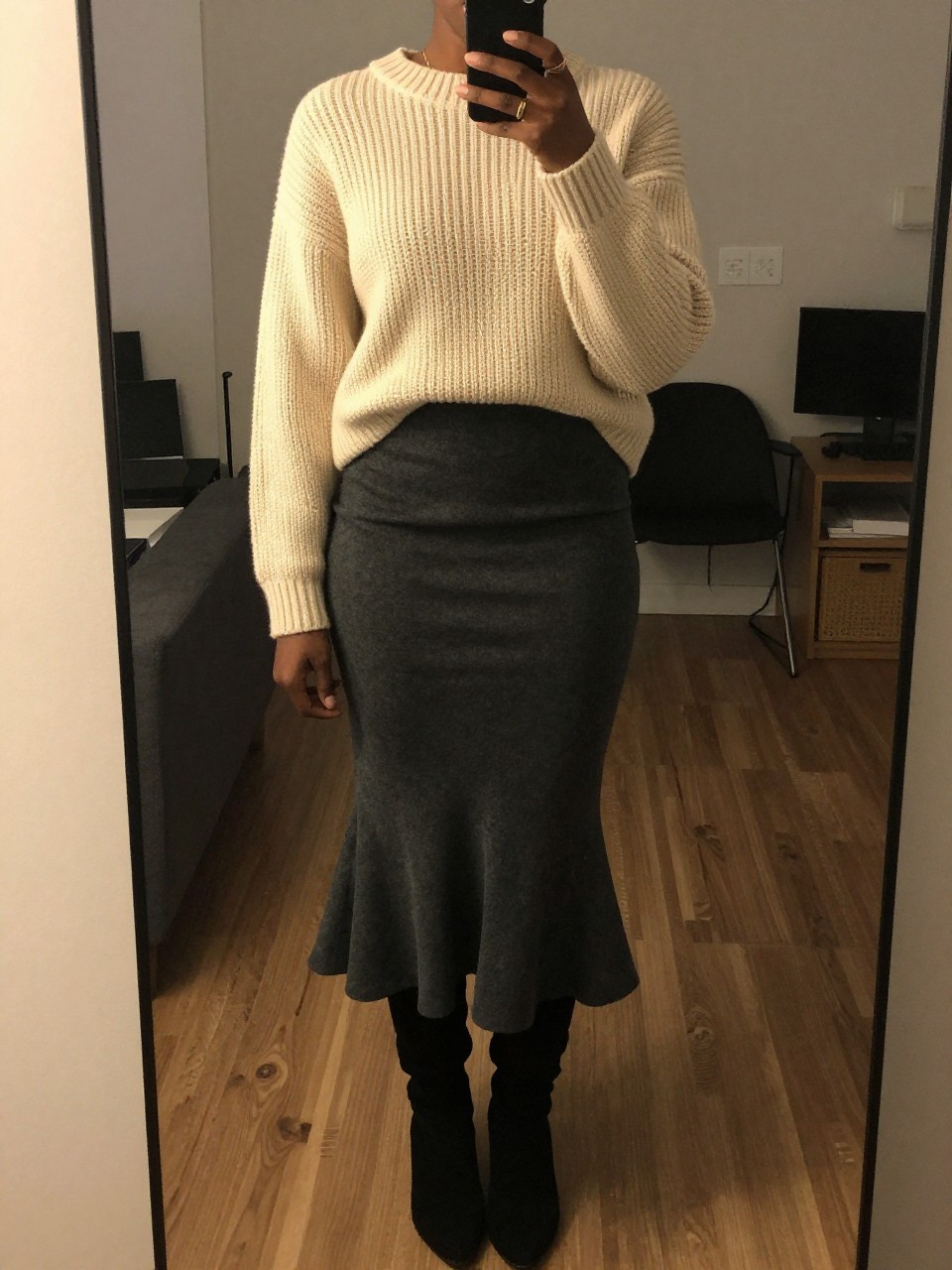 mirror selfie of a black woman in a cream chunky sweater and a charcoal mermaid skirt with knee-high boots, standing with phone in hand, cropped at NECK so face doesn't show, in a city apartment, warm moody lighting, iPhone photo quality.