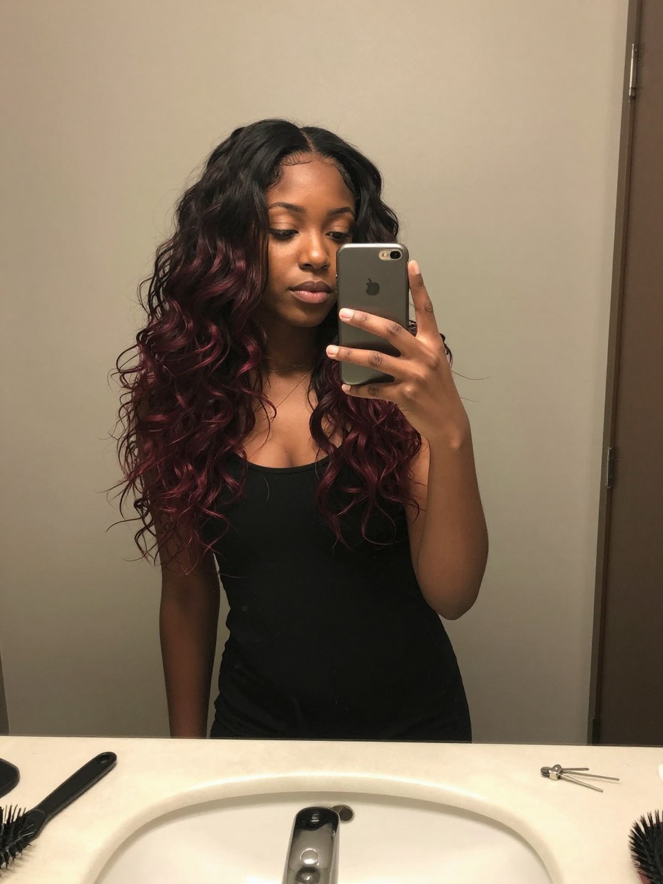 Photo of a black woman wearing a wavy black lob with dark red highlights, candid mirror selfie, at a bathroom vanity mirror, soft indoor lighting, containing a small lived-in detail such as a hairbrush on the sink and a silver clip nearby, latest iPhone photo quality.
