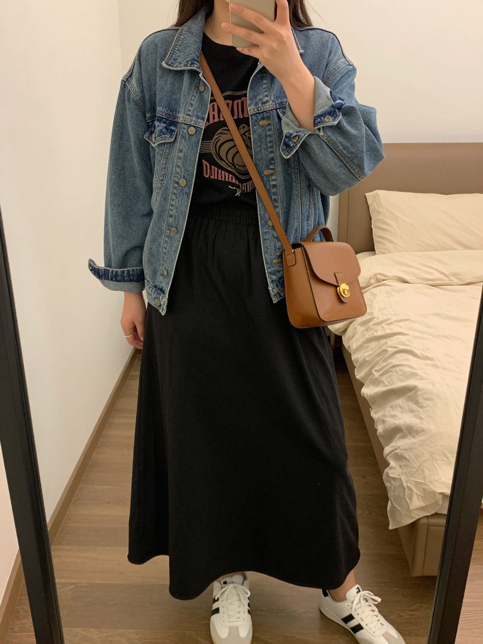 mirror selfie of a curvy woman wearing a black vintage band tee tucked into a black cotton maxi skirt with a light-wash denim jacket worn open and white sneakers, holding a tan leather crossbody bag, standing in a casual classic pose, cropped at NECK so face doesn't show, in a feminine-themed bedroom, warm moody lighting, iPhone photo quality.