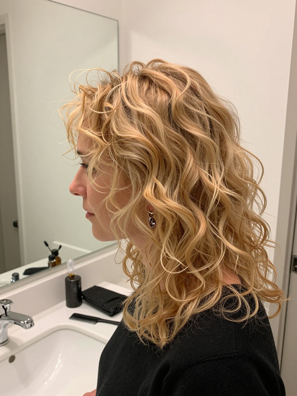 Photo of a woman wearing strawberry blonde curly layers with soft volume and defined curls, side profile view, bathroom vanity mirror setting, soft indoor lighting, with a wide-tooth comb on the counter and statement earrings peeking through, latest iPhone photo quality.