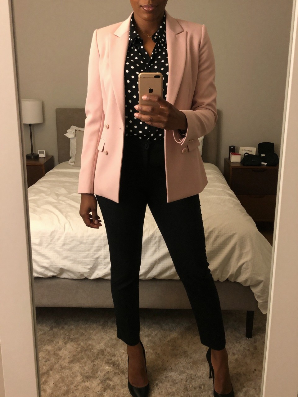 mirror selfie of a black woman wearing a pink blazer over a polka dot blouse with black trousers and heels, standing with phone near waist, cropped at NECK so face doesn't show, in a feminine-themed bedroom, warm moody lighting, iPhone photo quality.