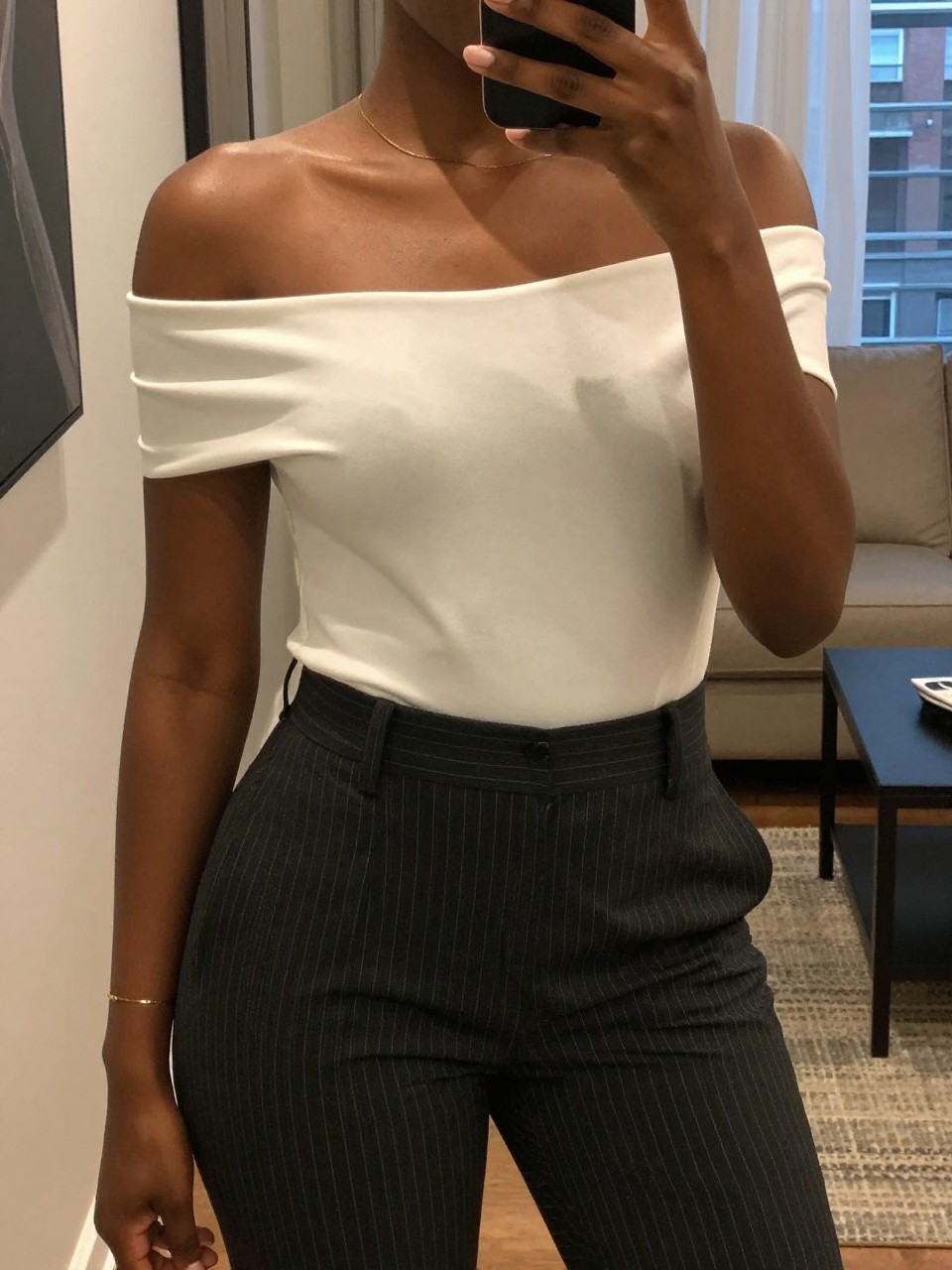 mirror selfie of a black woman in an off-the-shoulder top and tuxedo stripe pants, showing off her neckline, cropped at NECK so face doesn't show, in a city apartment, warm moody lighting, iPhone photo quality.