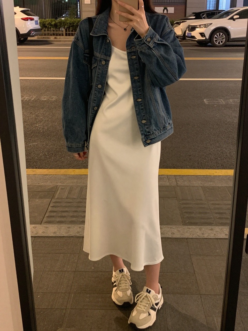 mirror selfie of a woman in a white slip dress and denim jacket with sneakers, standing on a city street or boardwalk, cropped at NECK so face doesn't show, in a city apartment, warm moody lighting, iPhone photo quality.