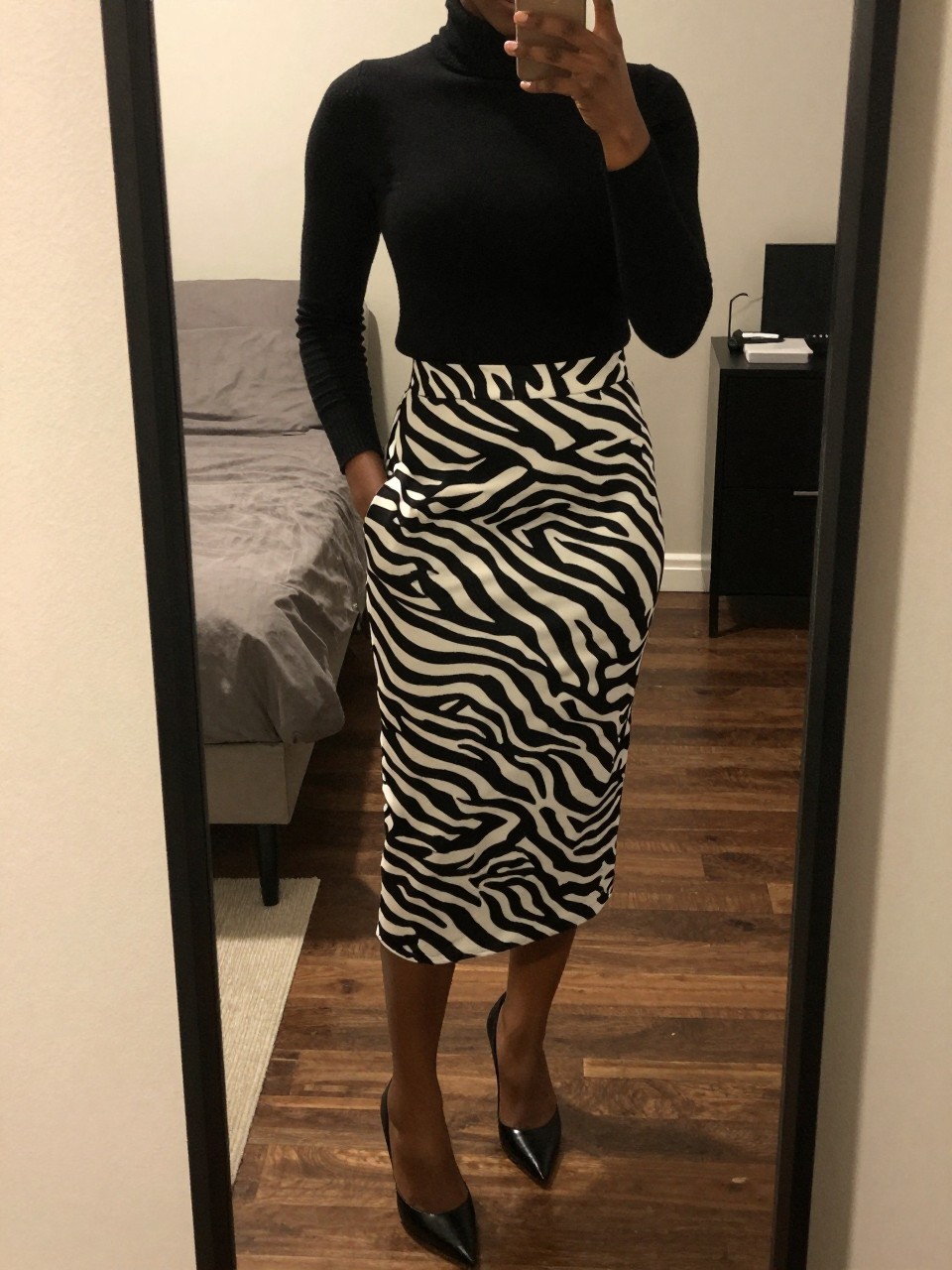 mirror selfie of a black woman in a black turtleneck and a zebra print midi skirt with pointed heels, standing with one hand in her pocket, cropped at NECK so face doesn't show, in a city apartment, warm moody lighting, iPhone photo quality.