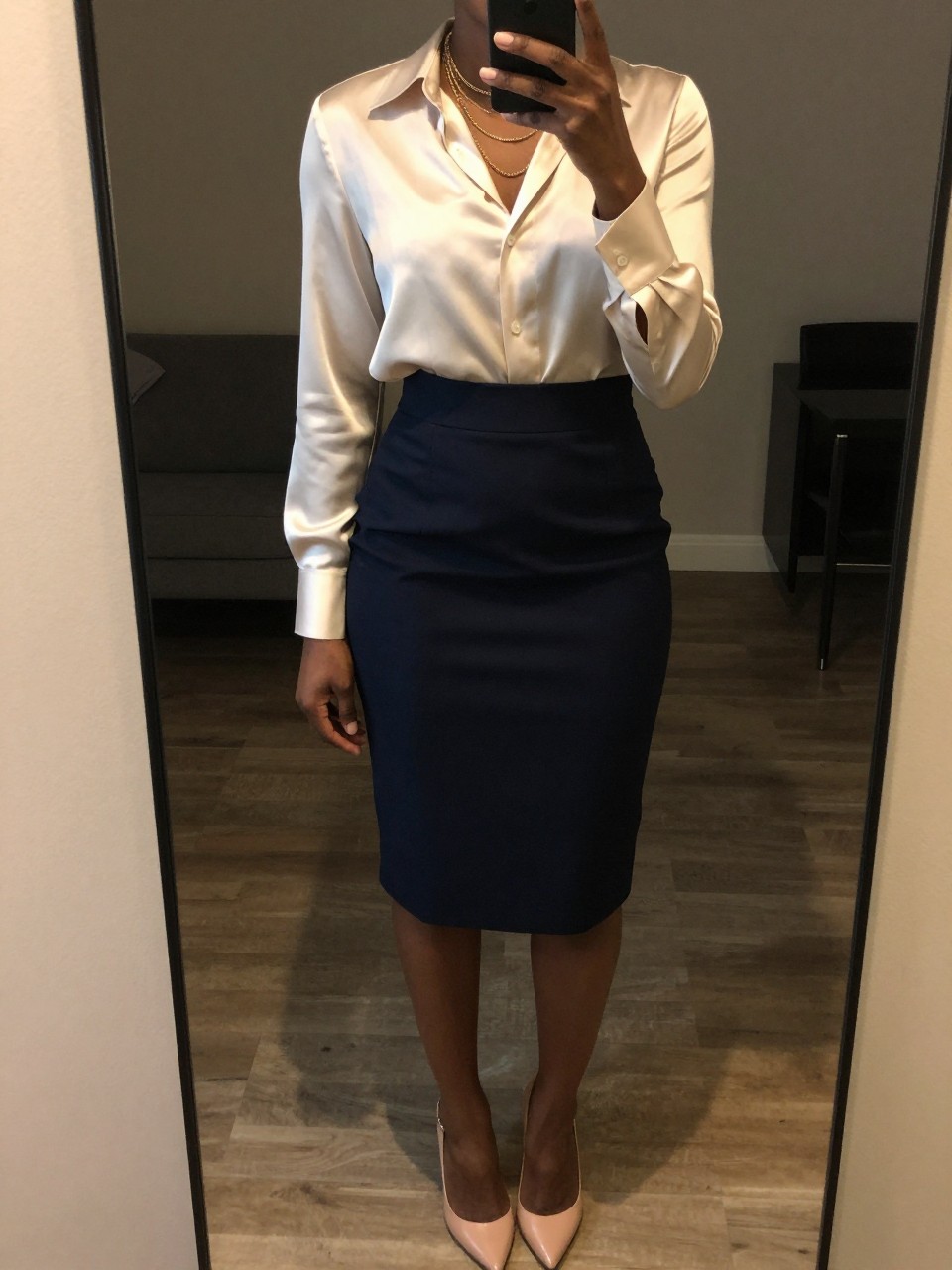 iPhone mirror selfie of a black woman wearing a cream silk blouse tucked into a navy wool pencil skirt with layered gold necklaces and nude pumps, one hand holding her phone at her side, cropped at NECK so face doesn't show, in a city apartment, warm moody lighting, iPhone photo quality.
