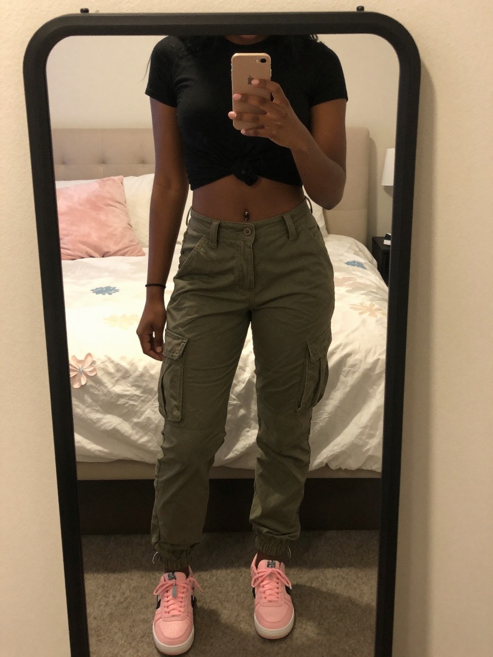 iPhone mirror selfie of a black woman in olive cargo pants and a black crop top with pink sneakers, holding phone at chest, cropped at NECK so face doesn't show, in a feminine-themed bedroom, warm moody lighting, iPhone photo quality.
