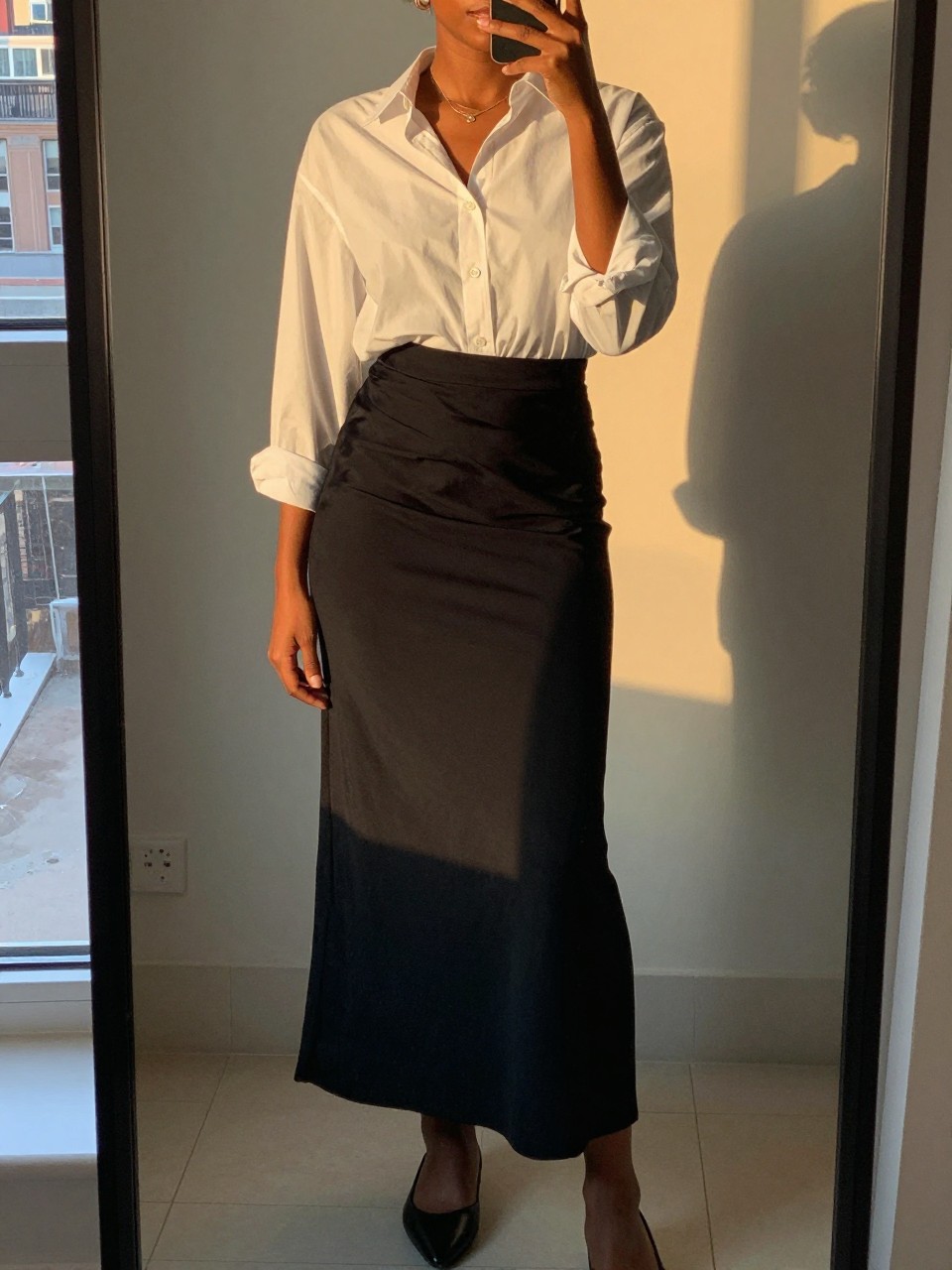 mirror selfie of a black woman in a black maxi skirt and white button-down with flats, standing with weight shifted, cropped at NECK so face doesn't show, in a city apartment, evening golden hour, iPhone photo quality.