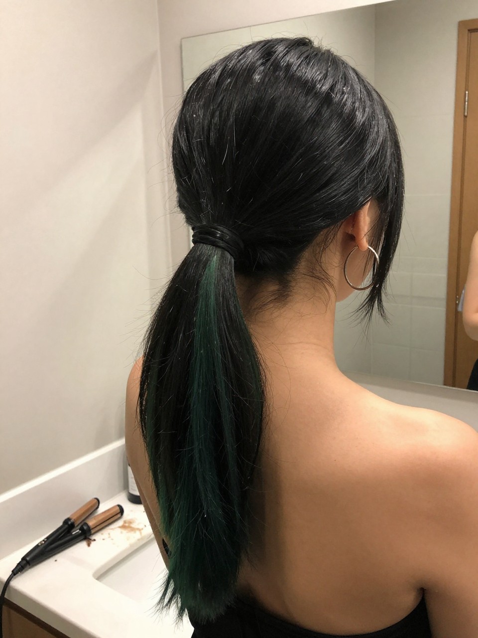 Photo of a woman wearing a sleek mid ponytail with black hair and green highlight panels through the tail, back-of-head view to highlight details, at a bathroom vanity mirror, soft indoor lighting, containing a small lived-in detail such as a flat iron on the counter and hoop earrings peeking through, latest iPhone photo quality.