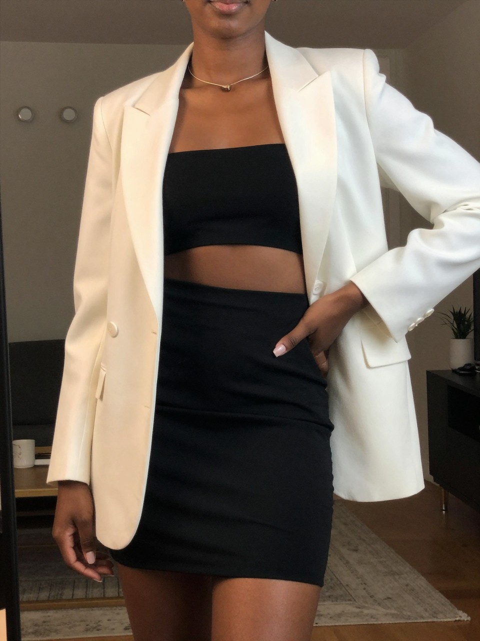 iPhone mirror selfie of a black woman in a cut-out black dress with a white blazer, hand on hip, cropped at NECK so face doesn't show, in a city apartment, warm moody lighting, iPhone photo quality.