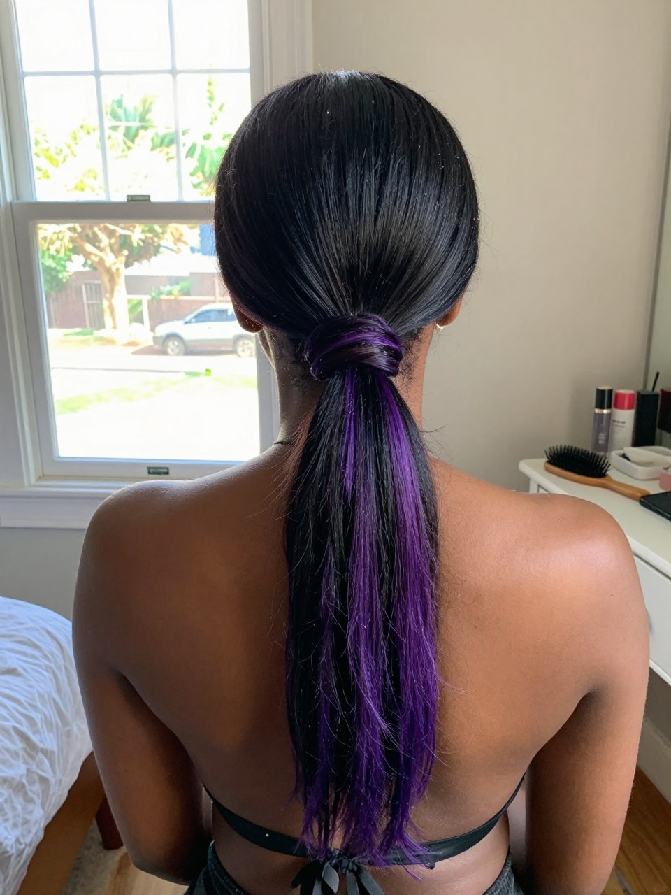 Photo of a black woman wearing a sleek low ponytail with black hair and purple highlights through the tail and ends, back-of-head view to highlight details, in a sun-drenched bedroom, natural window light, containing a small lived-in detail such as a black ribbon tied at the base and a hairbrush on the vanity, latest iPhone photo quality.