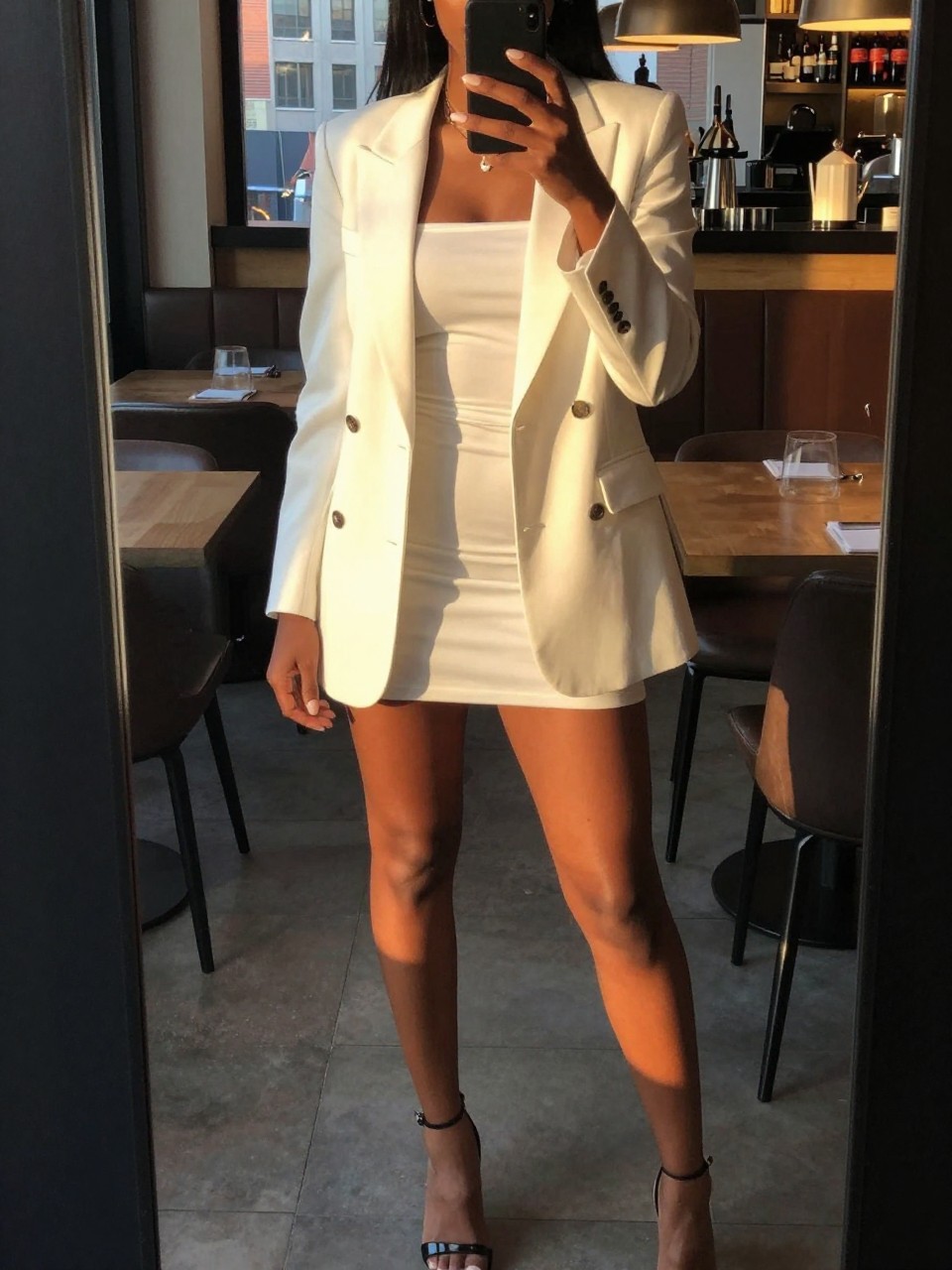 mirror selfie of a black woman in a white blazer over a white mini dress with heels, standing in a restaurant or bar, cropped at NECK so face doesn't show, in a city apartment, evening golden hour, iPhone photo quality.