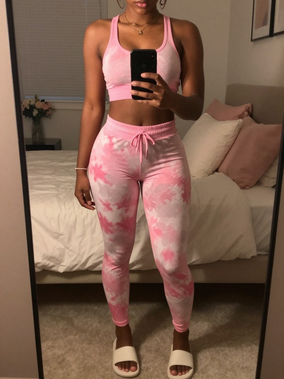 iPhone mirror selfie of a black woman in a pink tie-dye set with slides, standing with phone at waist, cropped at NECK so face doesn't show, in a feminine-themed bedroom, warm moody lighting, iPhone photo quality.