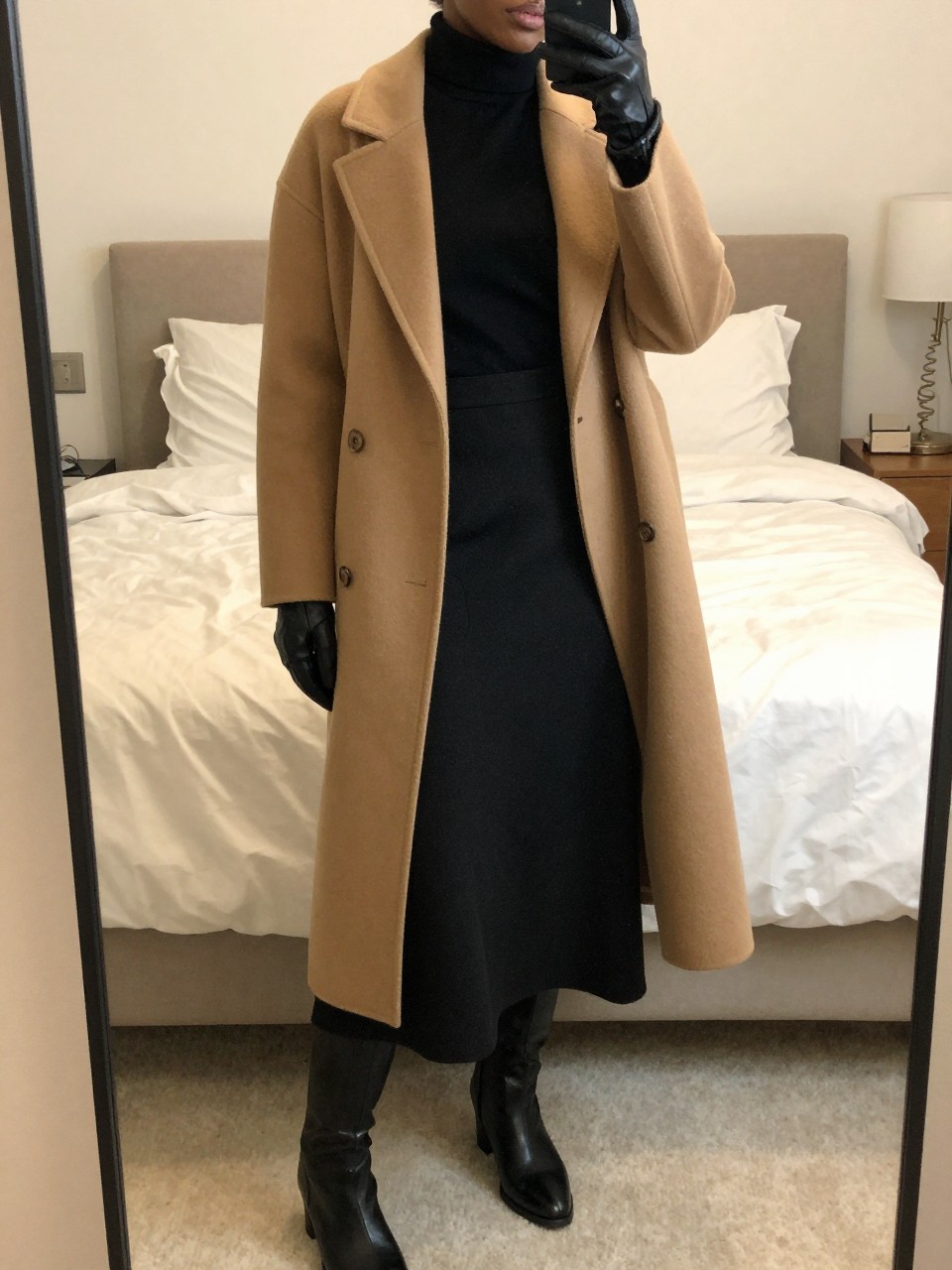 mirror selfie of a black woman wearing a black turtleneck tucked into a black wool maxi skirt with a long camel wool coat over top and black leather knee-high boots, holding leather gloves, standing in an elegant winter pose, cropped at NECK so face doesn't show, in a feminine-themed bedroom, warm moody lighting, iPhone photo quality.