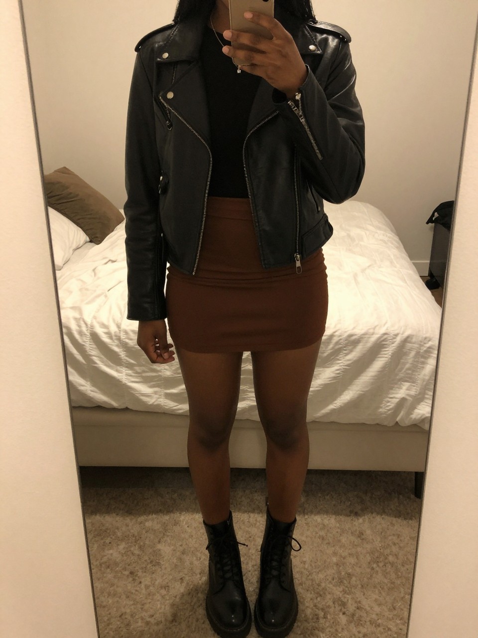 iPhone mirror selfie of a black woman wearing a black leather jacket over a band tee with a chocolate brown mini skirt and black combat boots, one hand in her jacket pocket, cropped at NECK so face doesn't show, in a feminine-themed bedroom, warm moody lighting, iPhone photo quality.