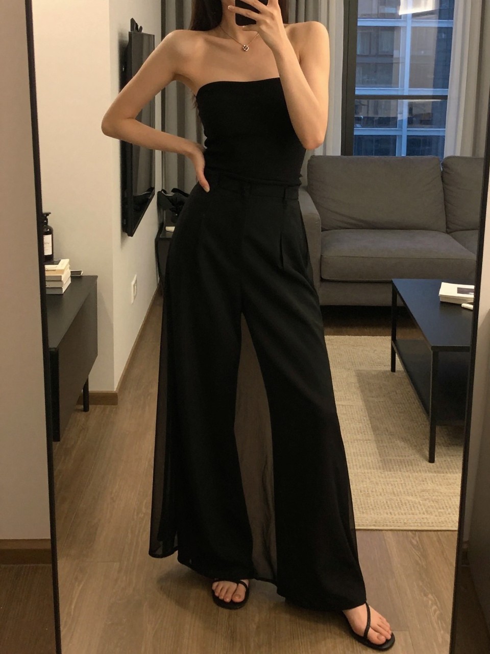 mirror selfie of a woman wearing a black bandeau top with black wide-leg trousers underneath a sheer black mesh maxi skirt and flat strappy sandals, one hand on her hip, cropped at NECK so face doesn't show, in a city apartment, warm moody lighting, iPhone photo quality.