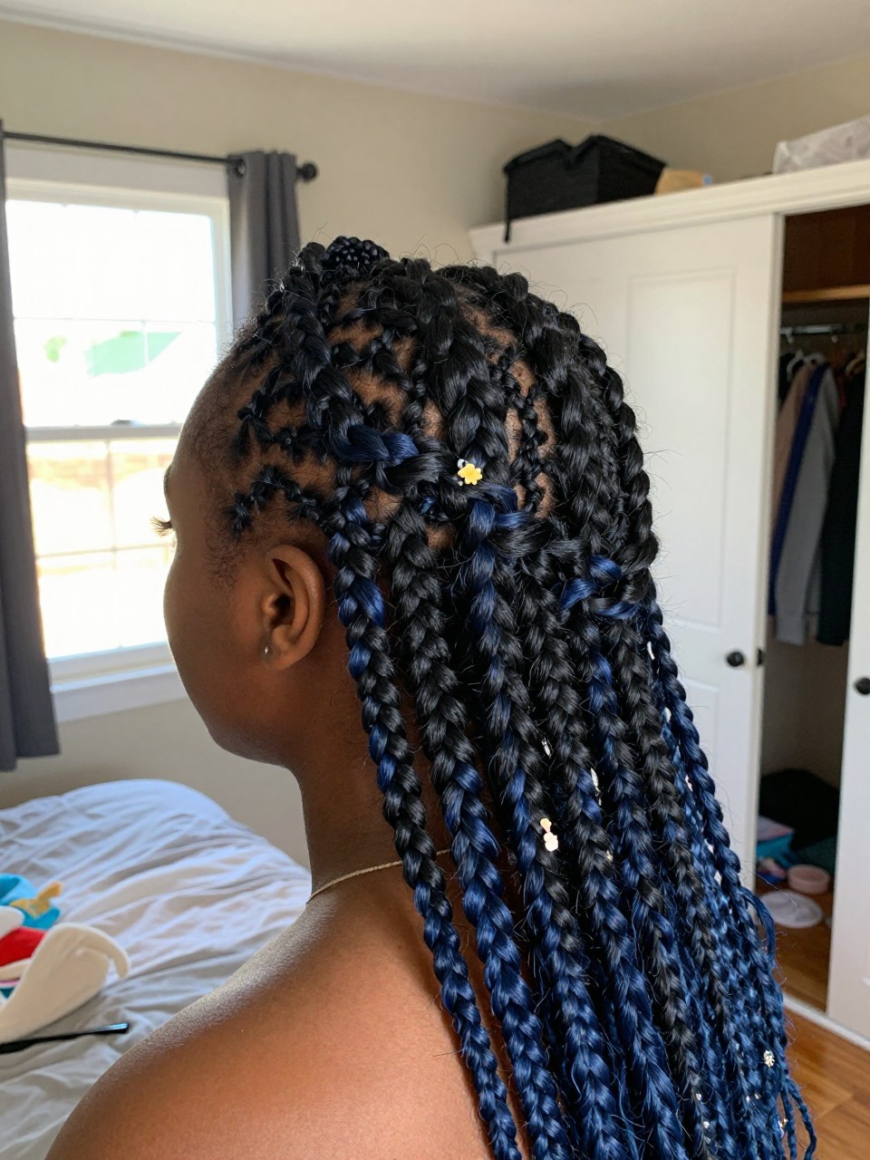 Photo of a black woman wearing a braided crown on black hair with navy blue highlights woven throughout, back-of-head view to highlight details, in a sun-drenched bedroom, natural window light, containing a small lived-in detail such as a tiny flower pin and a slightly open closet behind, latest iPhone photo quality.