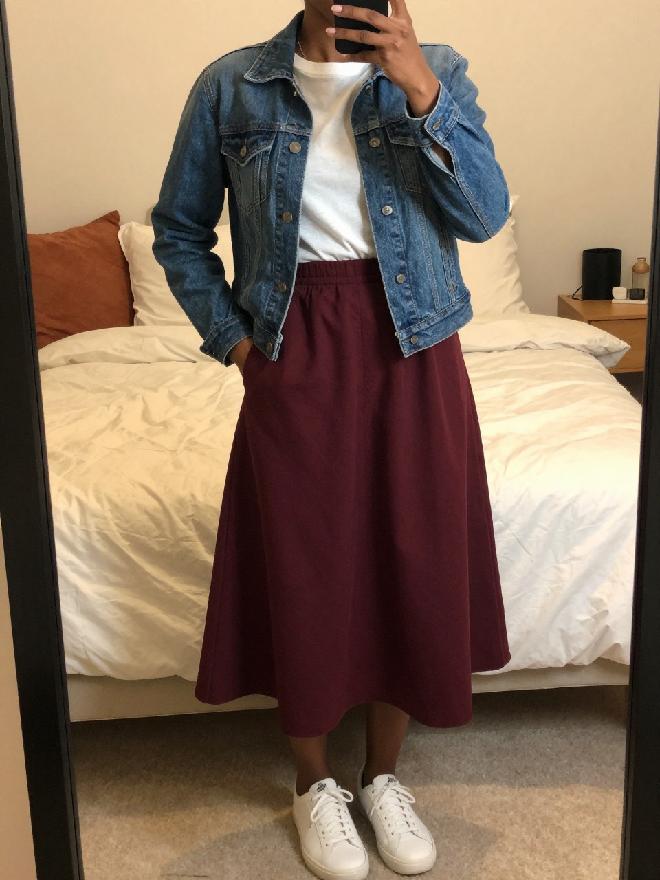 mirror selfie of a black woman wearing a blue denim jacket over a white tee tucked into a deep burgundy cotton A-line skirt with white sneakers, one hand in her jacket pocket, cropped at NECK so face doesn't show, in a feminine-themed bedroom, warm moody lighting, iPhone photo quality.