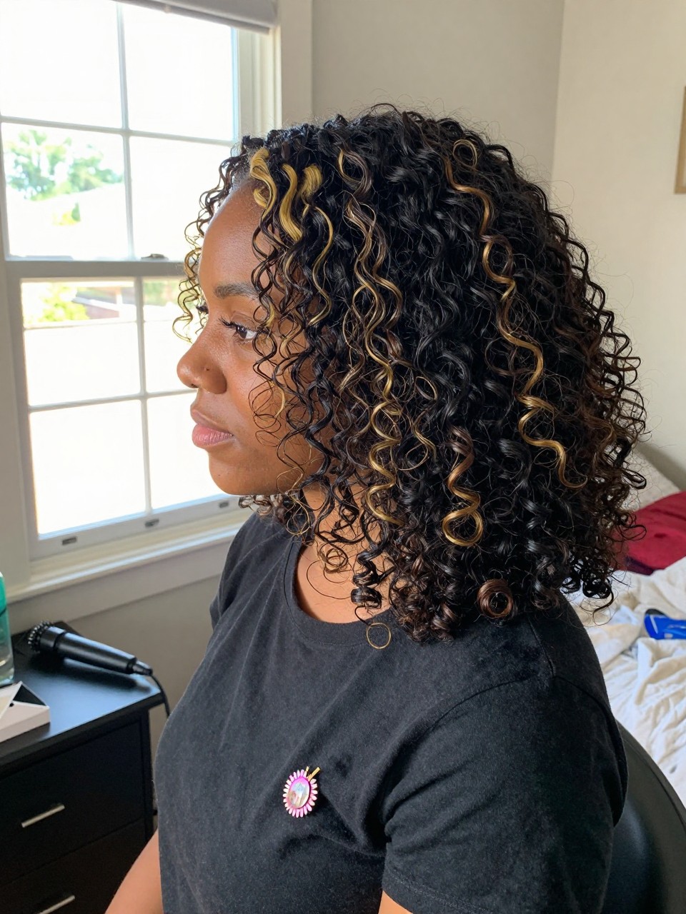 Photo of a black woman wearing soft black curls with golden blonde highlight streaks, side profile view, in a sun-drenched bedroom, natural window light, containing a small lived-in detail such as a decorative pin on one side and a curling iron resting on the nightstand, latest iPhone photo quality.