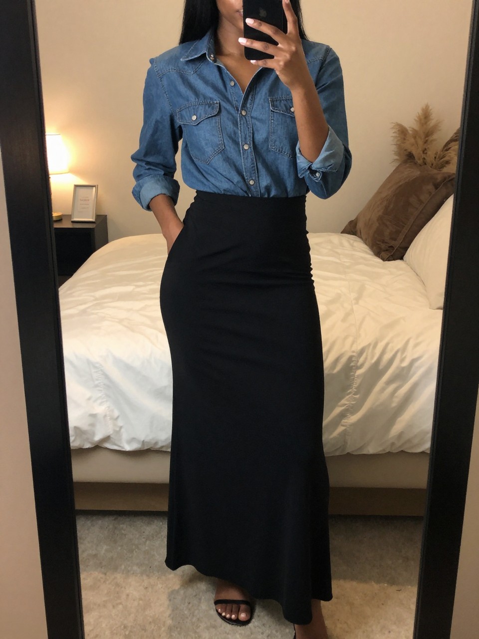 iPhone mirror selfie of a black woman in a black maxi skirt and denim shirt with sandals, standing with one hand in pocket, cropped at NECK so face doesn't show, in a feminine-themed bedroom, warm moody lighting, iPhone photo quality.