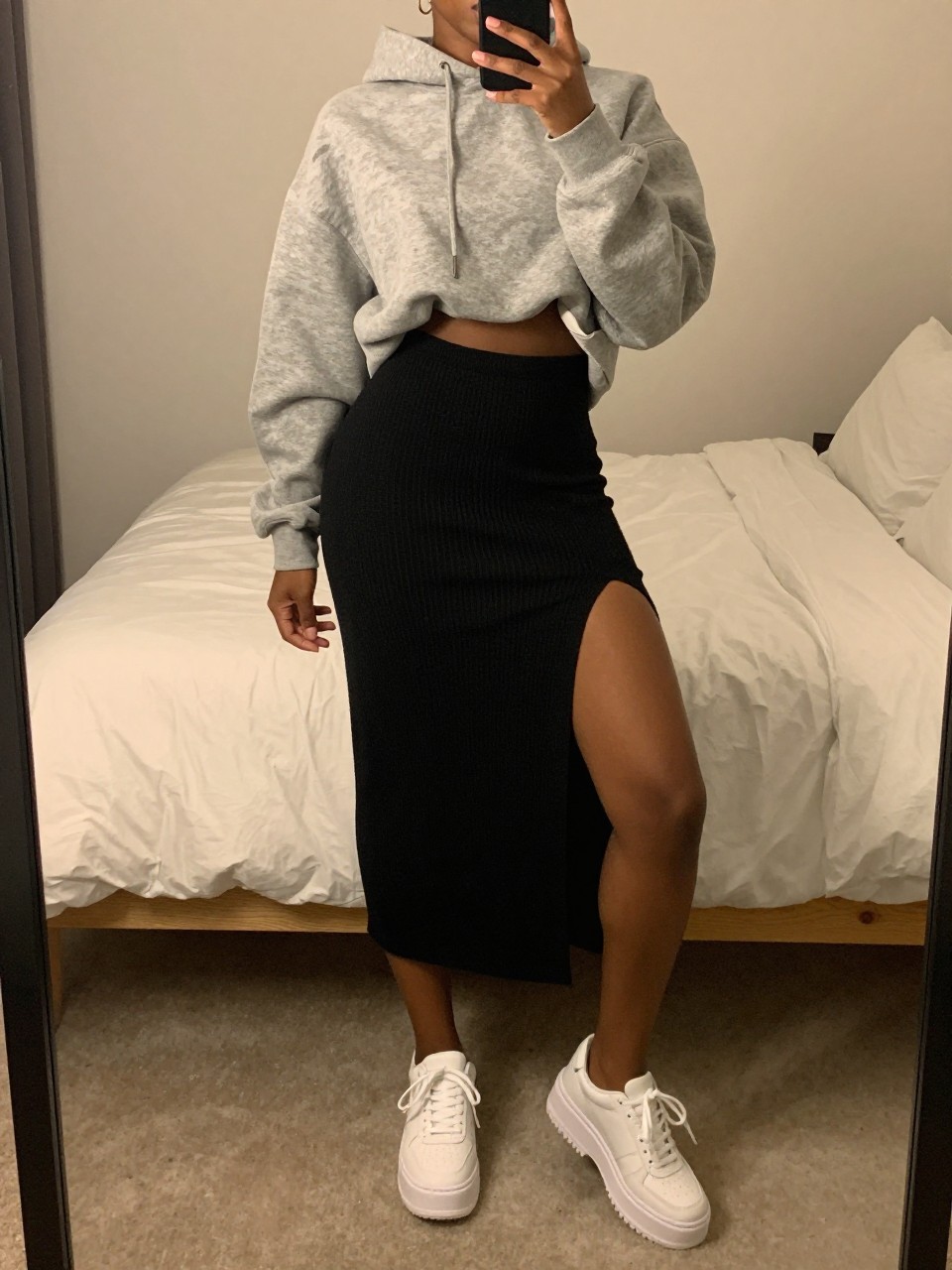mirror selfie of a black woman wearing an oversized gray hoodie half-tucked into a black knit midi skirt with chunky white platform sneakers, one hand holding her phone low by her thigh, cropped at NECK so face doesn't show, in a feminine-themed bedroom, warm moody lighting, iPhone photo quality.