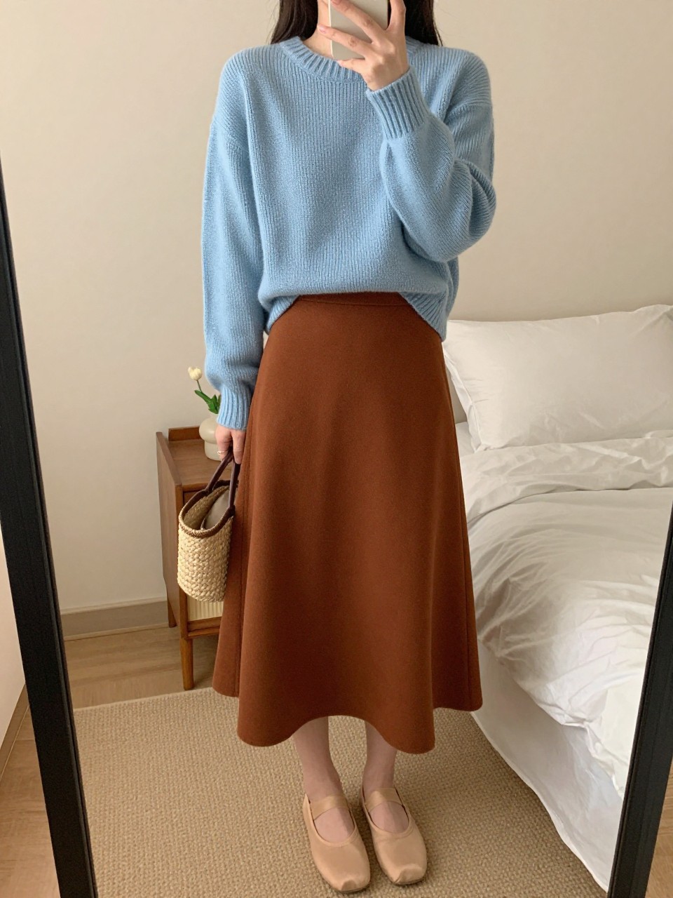 mirror selfie of a woman wearing a powder blue fine-knit sweater tucked into a rust brown A-line skirt with nude ballet flats and a woven straw bag, standing in a soft, feminine pose, cropped at NECK so face doesn't show, in a feminine-themed bedroom, warm moody lighting, iPhone photo quality.